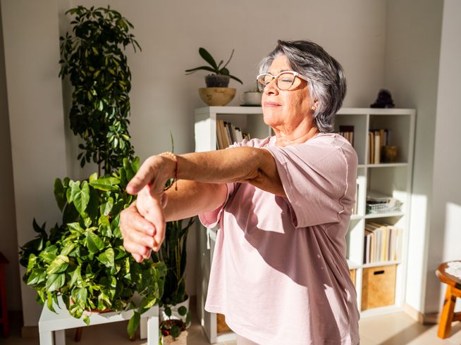 Senior woman stretching her left arm and hand while standing in the sunlight.