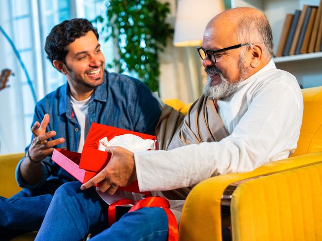 Adult son gifting senior father a gift on a yellow couch in a living room.